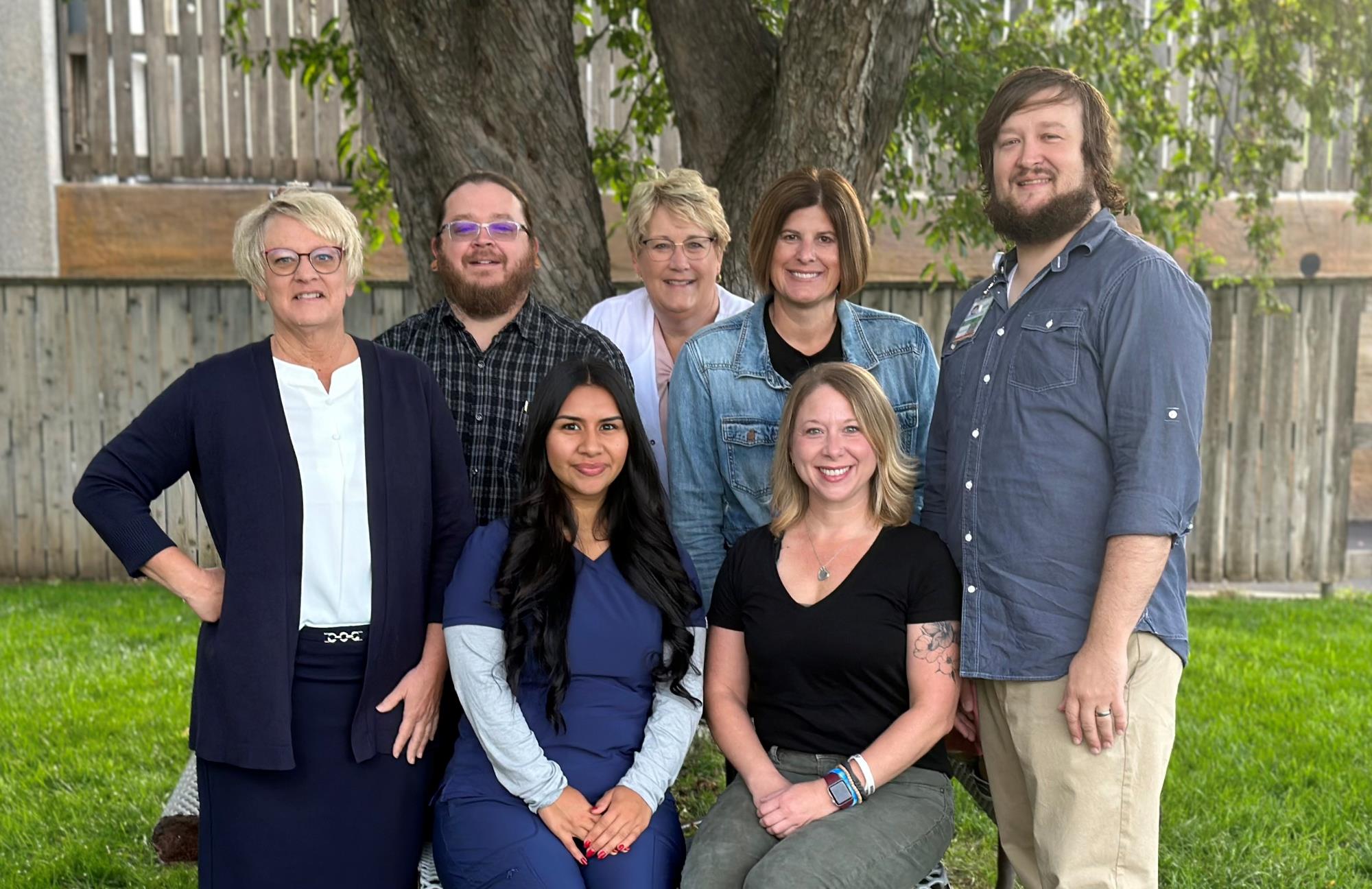 Group of seven SDHD employees standing outside in front of a tree smiling and looking straight ahead. 