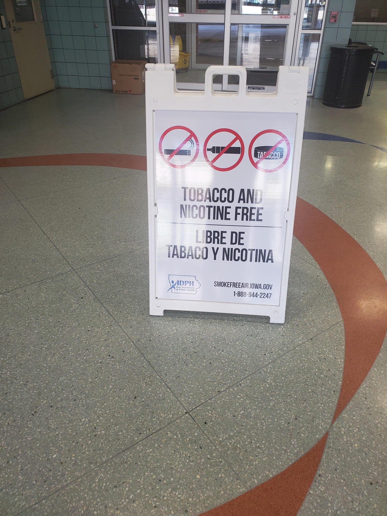 Sandwich board Tobacco and Nicotine Free Sign sitting on the ground in a lobby with automatic doors behind it.  Smokefreeair.iowa.gov 1-888-944-2247