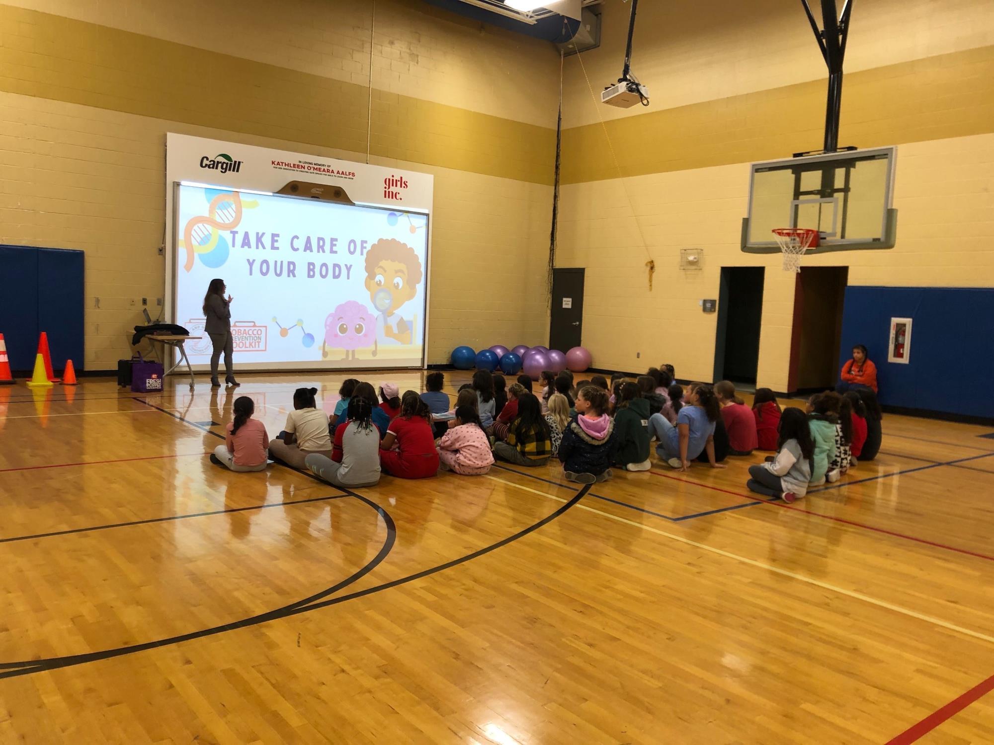 Adult female standing next to a screen in front of a classroom of kids who are sitting on a gym floor. Teacher sitting in a chair on the side.