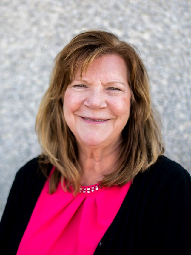 Woman with brown shoulder length hair, pink shirt and black sweater smiling