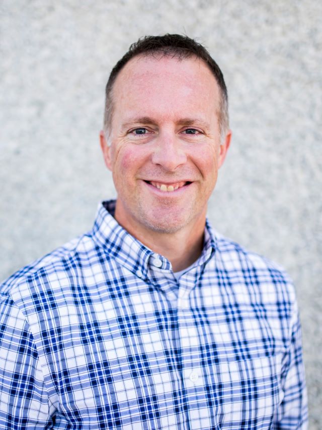 Man with short dark hair and blue and white collared shirt on smiling