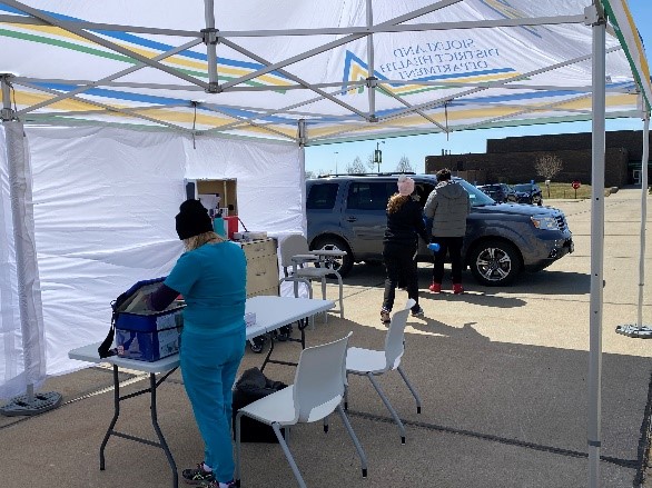 Person in blue scrubs at a table under a tent. Vehicle next to tent with two people near vehicle. 