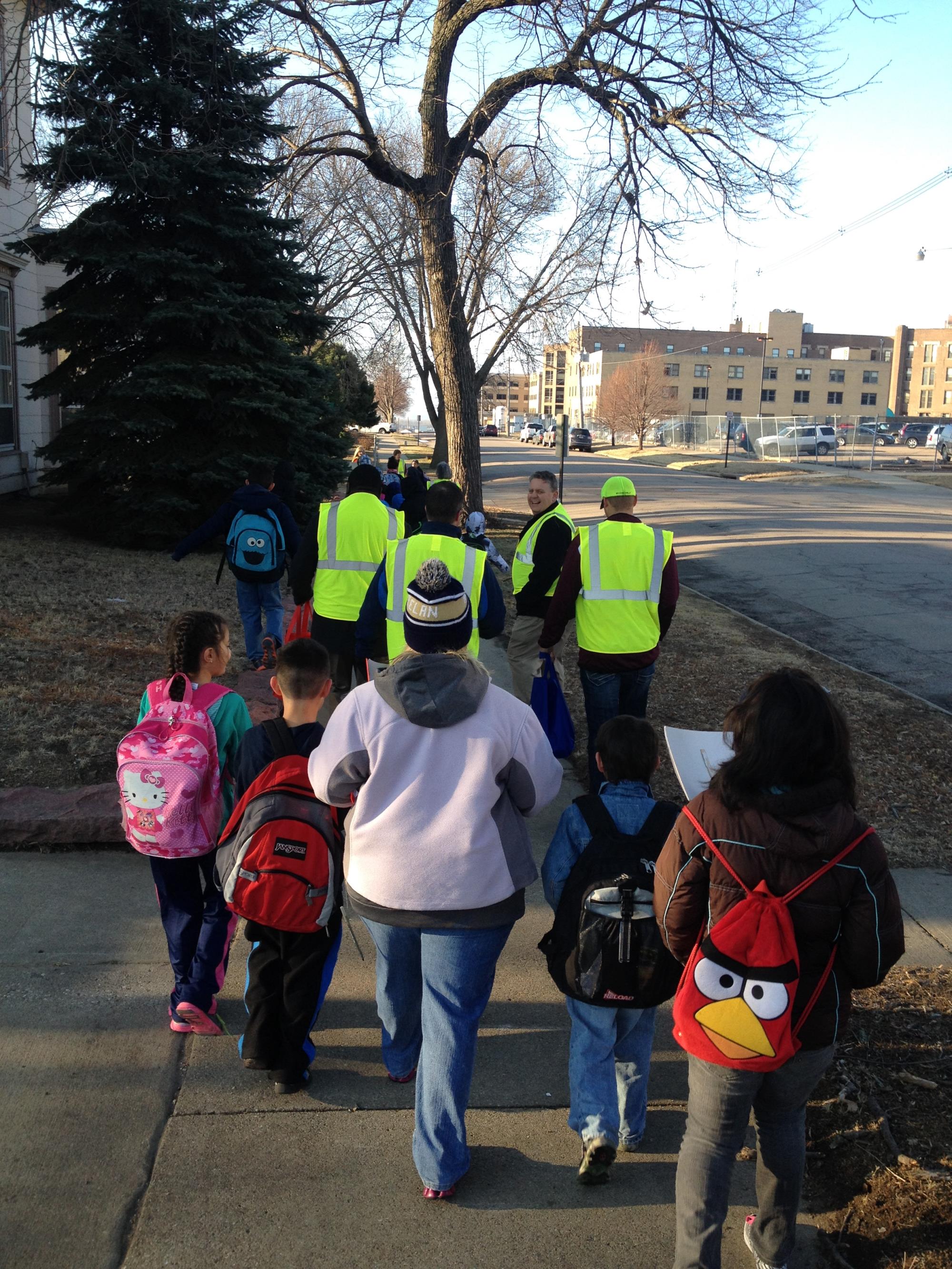 Adults with yellow vests walking with students with backpacks on. They are walking down a sidewalk. 