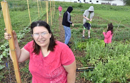 Person with pink shirt standing in a community garden. She is smiling looking at the camera. Four people are working in the garden behind her.