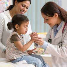 child sitting on table with person behind watching a female with lab coat on looking at and holding fingers on the child's right hand.