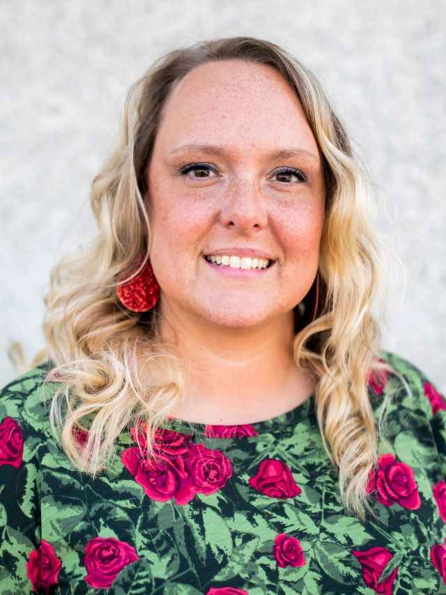 woman with longer curled blonde hair, pink roses and green leaves on shirt, smiling